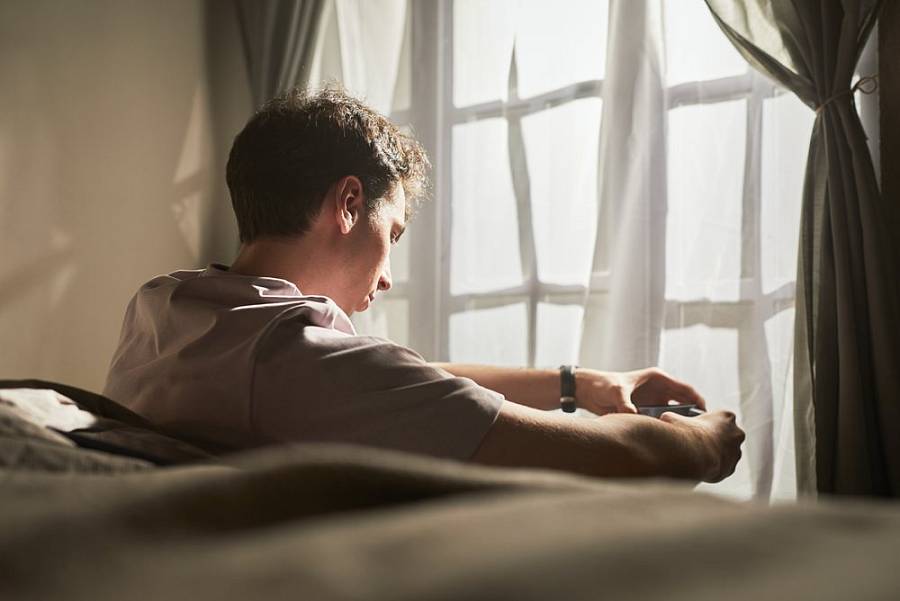 Nachdenkliche Person sitzt morgens am Fenster und blickt mit gesenktem Kopf auf ihre Hände.